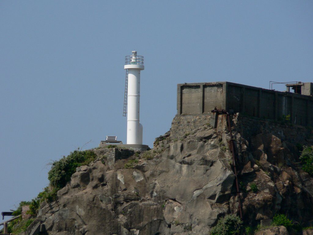 The Hashima lighthouse on a rocky outcrop, with a decaying concrete building visible behind it.